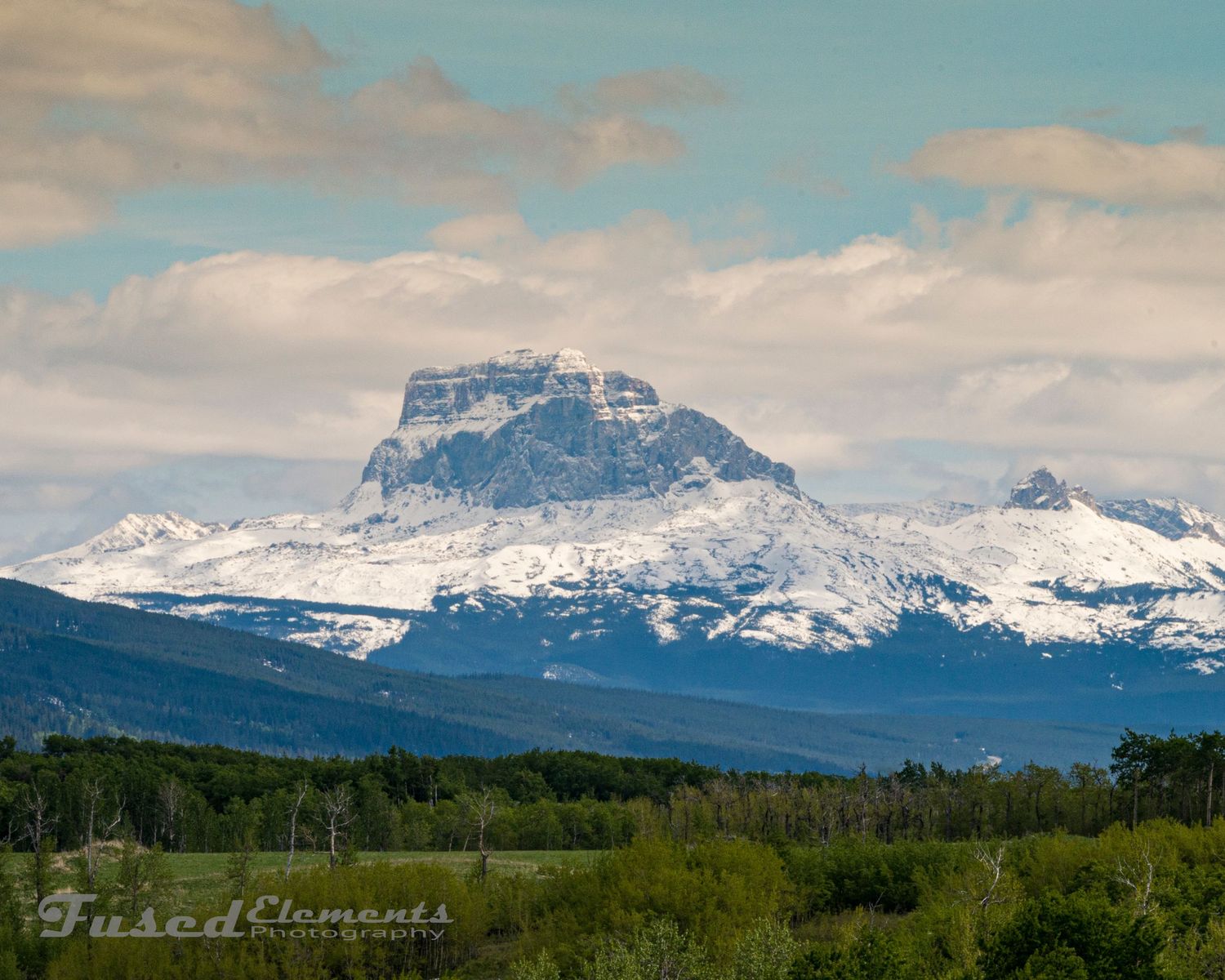 Snow-capped mountain peak rises majestically above rolling green foothills and forests in a dramatic landscape vista.