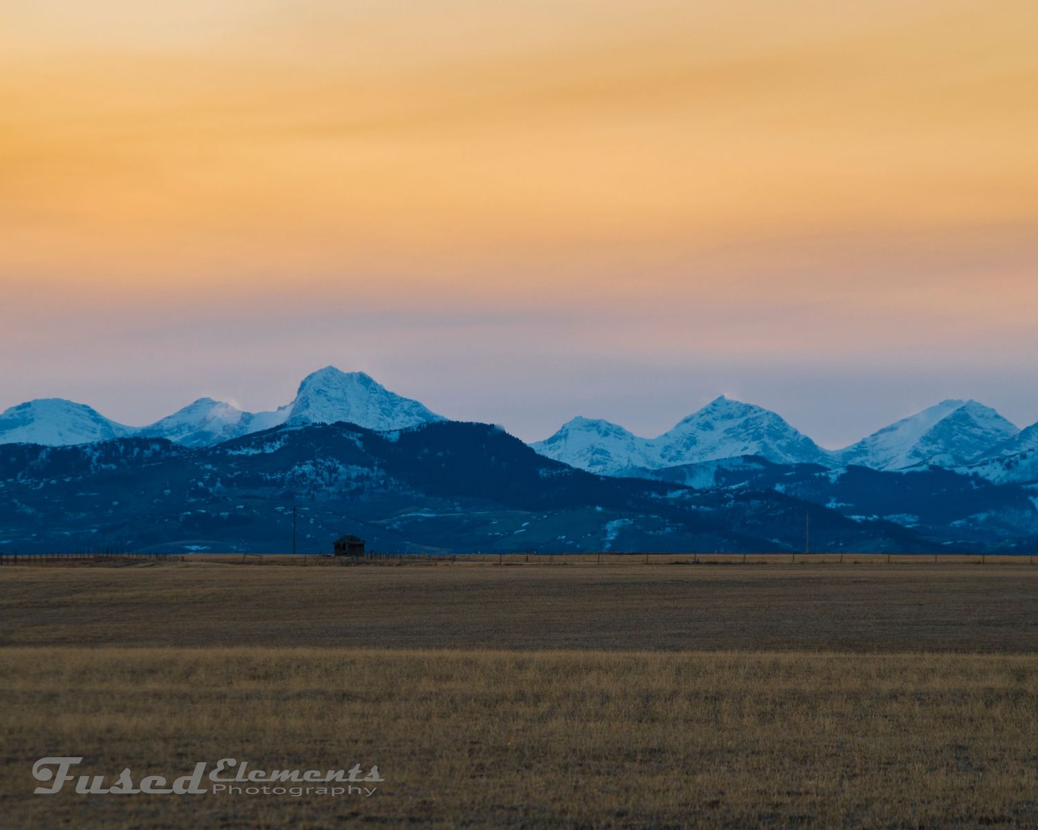 Snow-capped mountain range silhouetted against a golden sunset sky over open prairie.