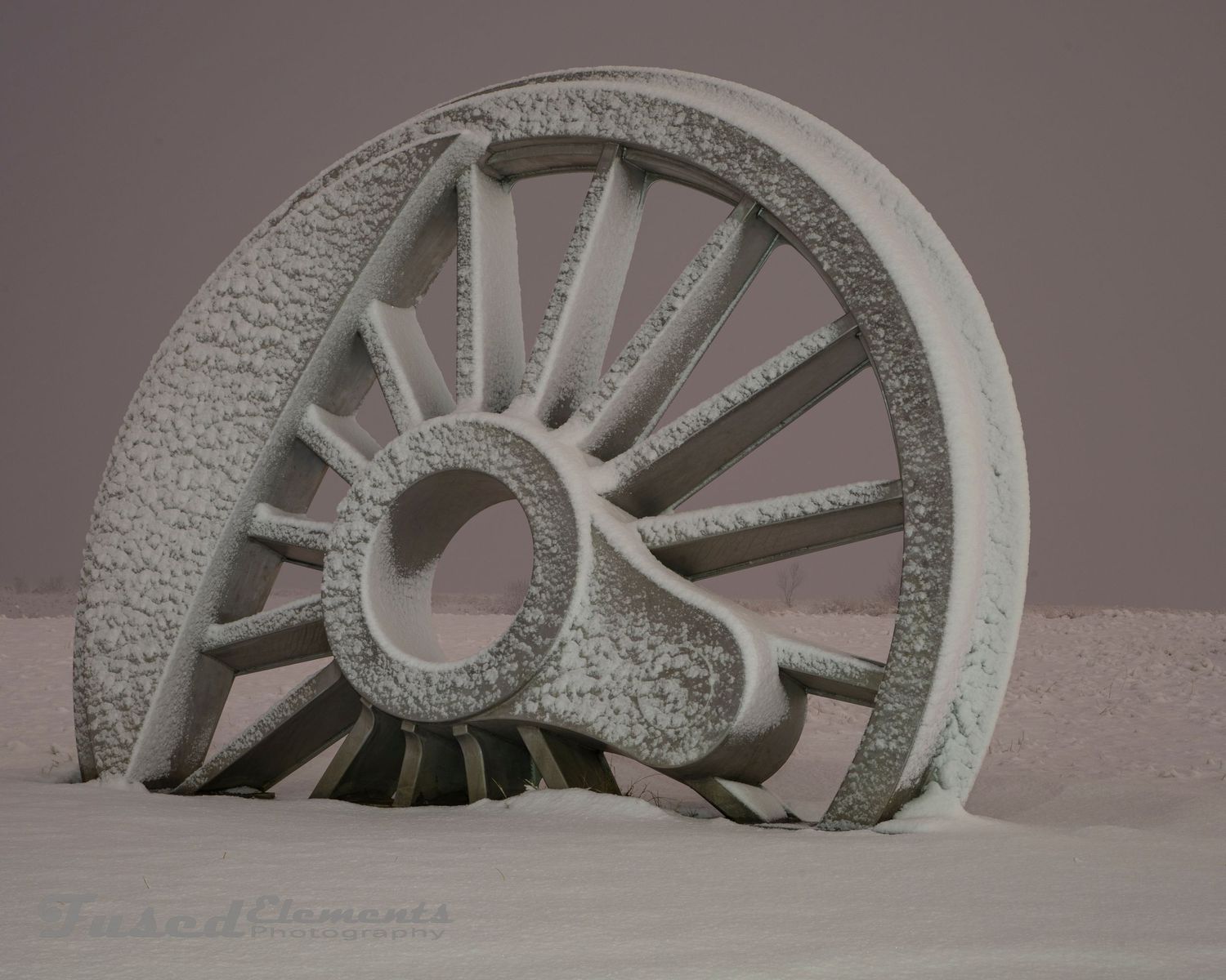Snow-covered sculptural wheel fragment creates abstract geometric shapes against muted background.
