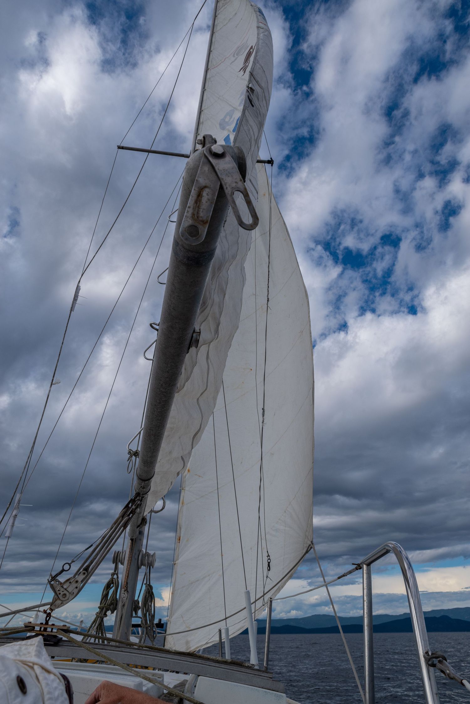A sailboat's white canvas sail and mast against a dramatic cloudy sky over water.