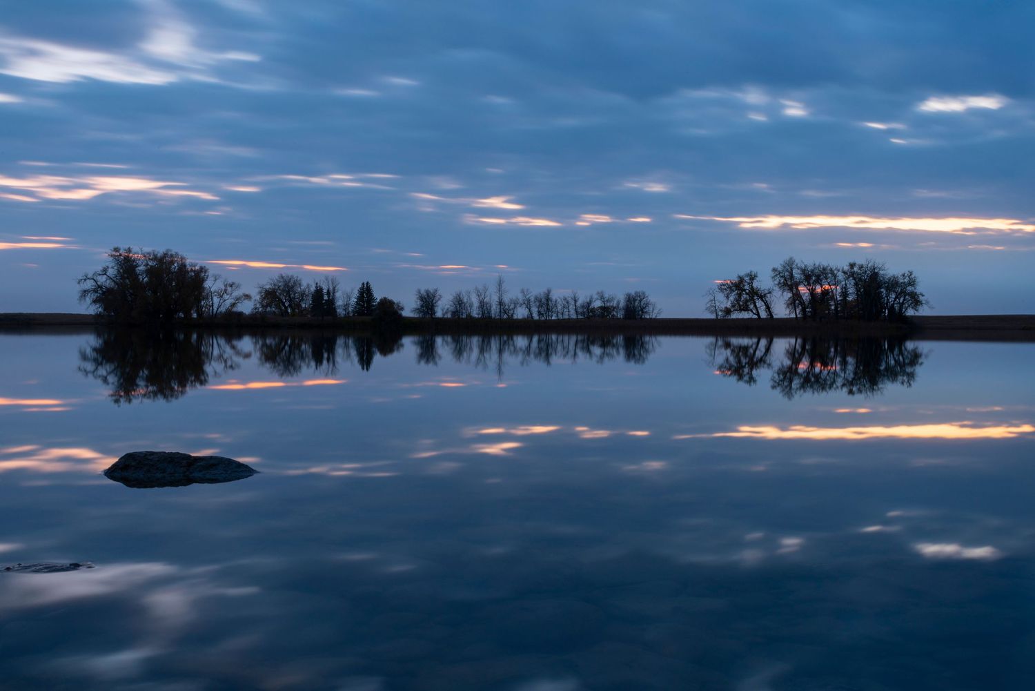 Tranquil lake reflects twilight sky and silhouetted treeline in perfect symmetry.