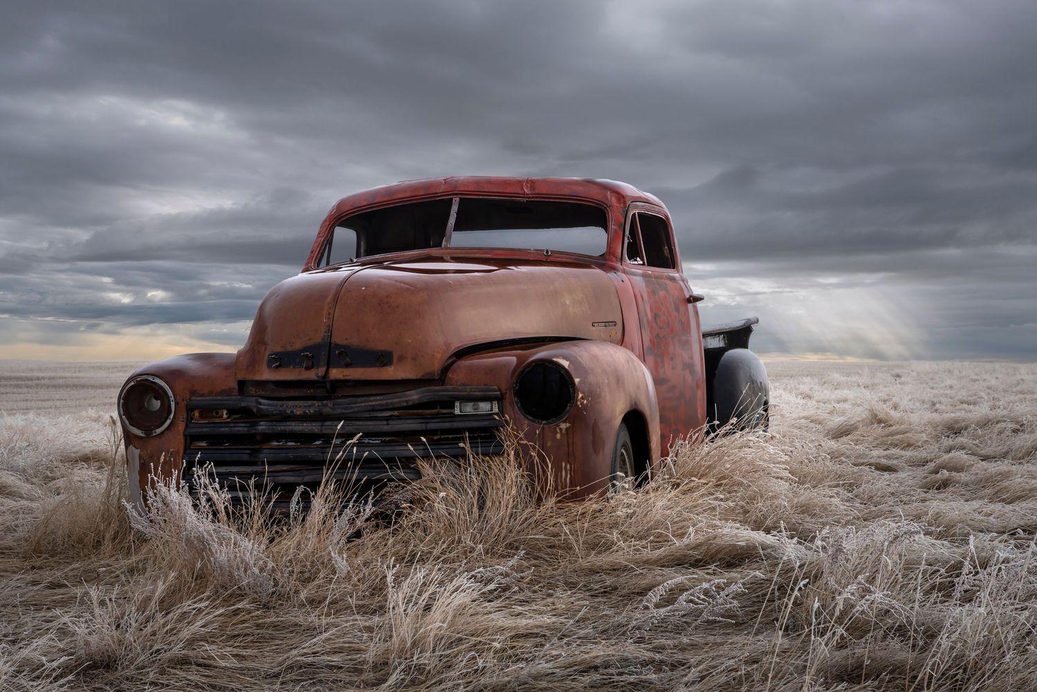 Rusty vintage Chevrolet truck abandoned in dry grass field under dramatic stormy sky.