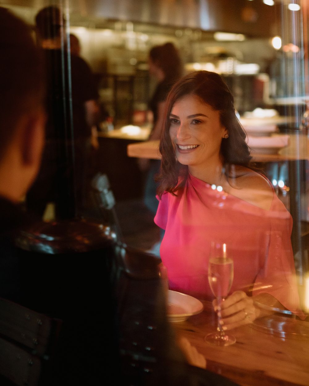 Woman in pink dress smiles at bar with champagne glass in romantic setting.