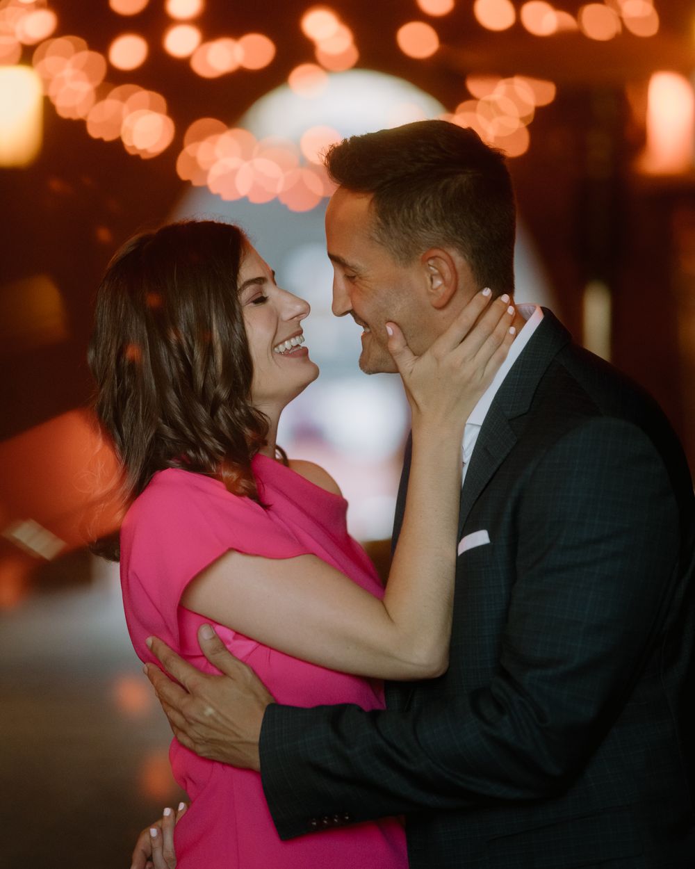 Intimate moment between couple in pink dress and dark suit with bokeh lighting.
