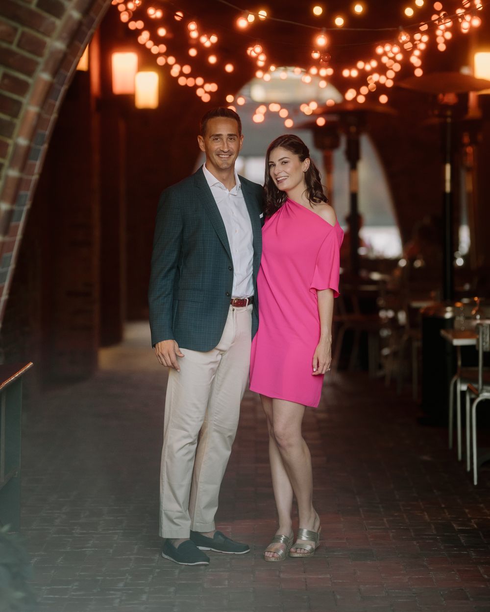 Couple in pink dress and green blazer poses under archway with string lights.