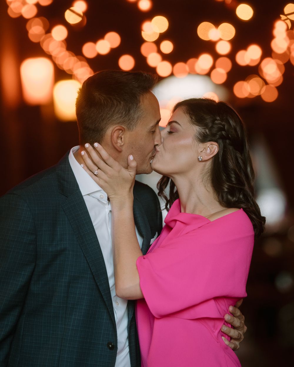 Romantic kiss between couple dressed in pink and dark suit with bokeh lights in background.