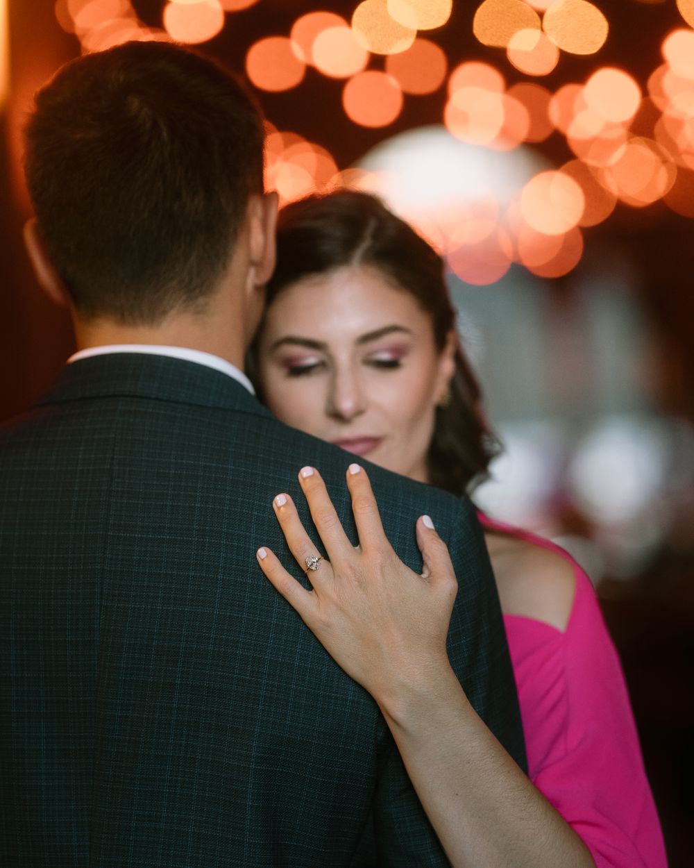Hand rests on dark suit jacket against bokeh lights backdrop in romantic setting.