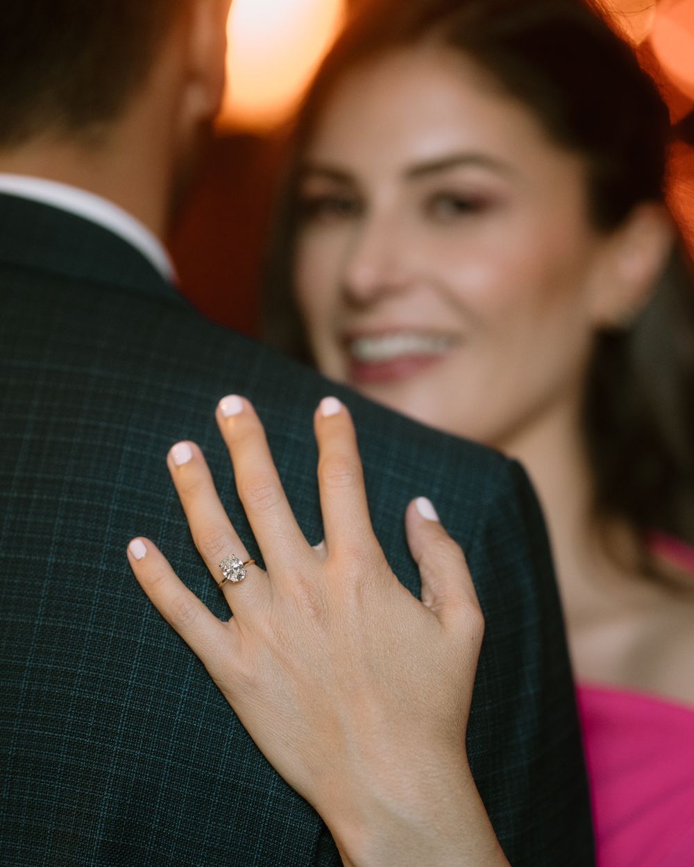 Woman's hand showing engagement ring while embracing partner in dark suit against warm lighting.