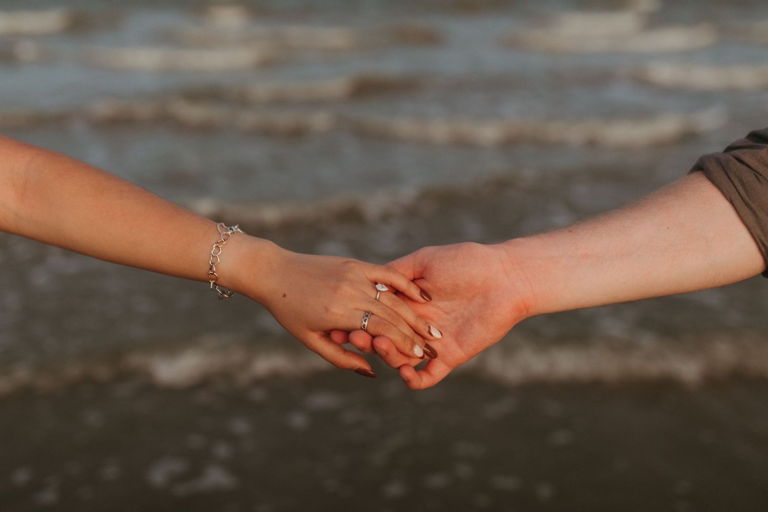 Close-up of two hands clasping together on a beach with ocean waves in the background.