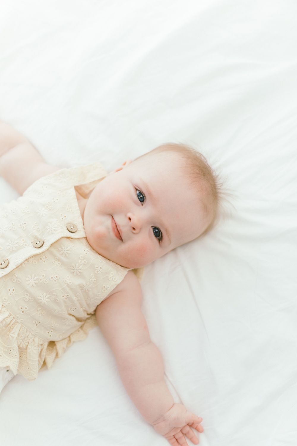 Baby in beige romper resting on white bedding looking at camera.