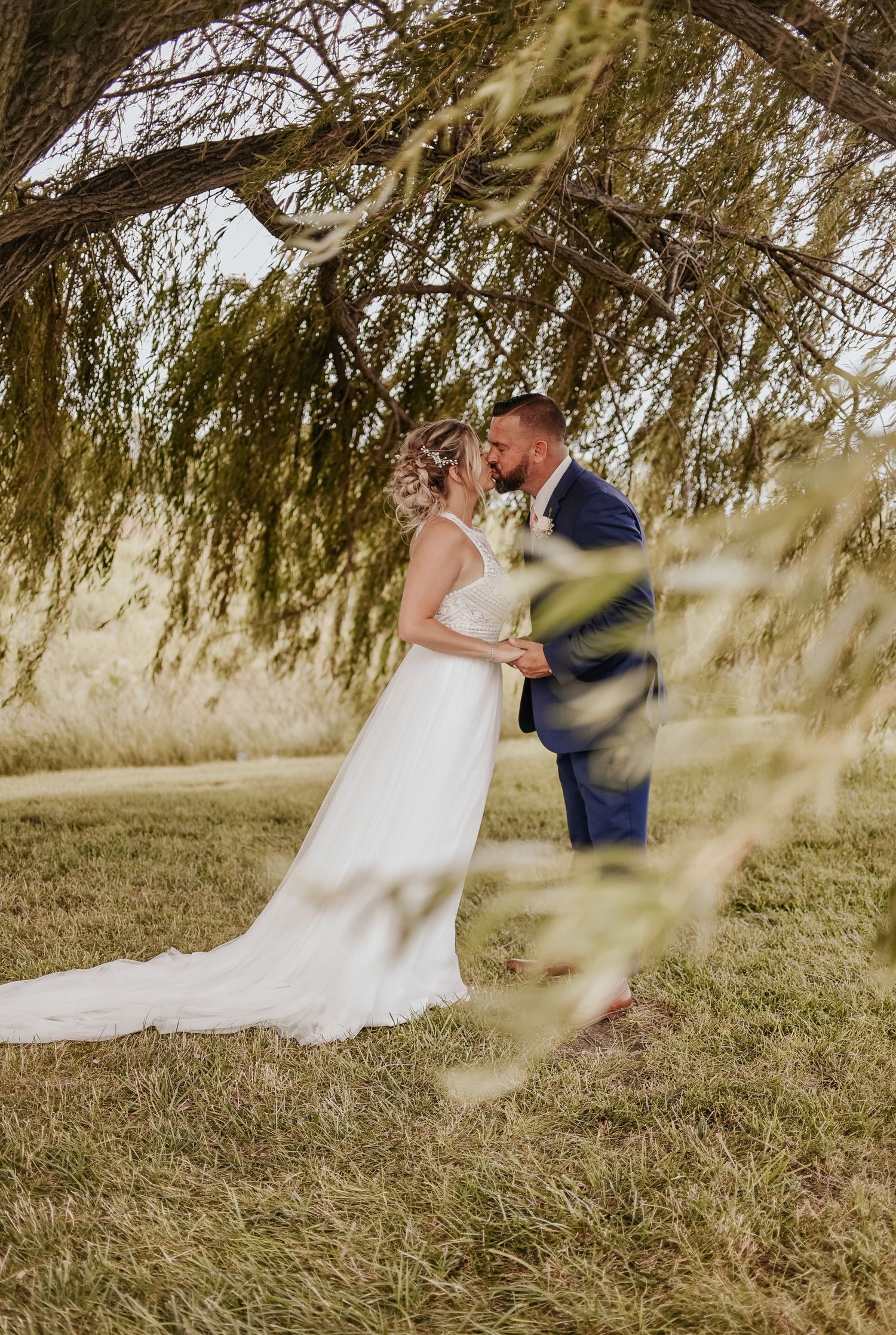 Wedding couple embracing under willow tree branches in golden sunlight.