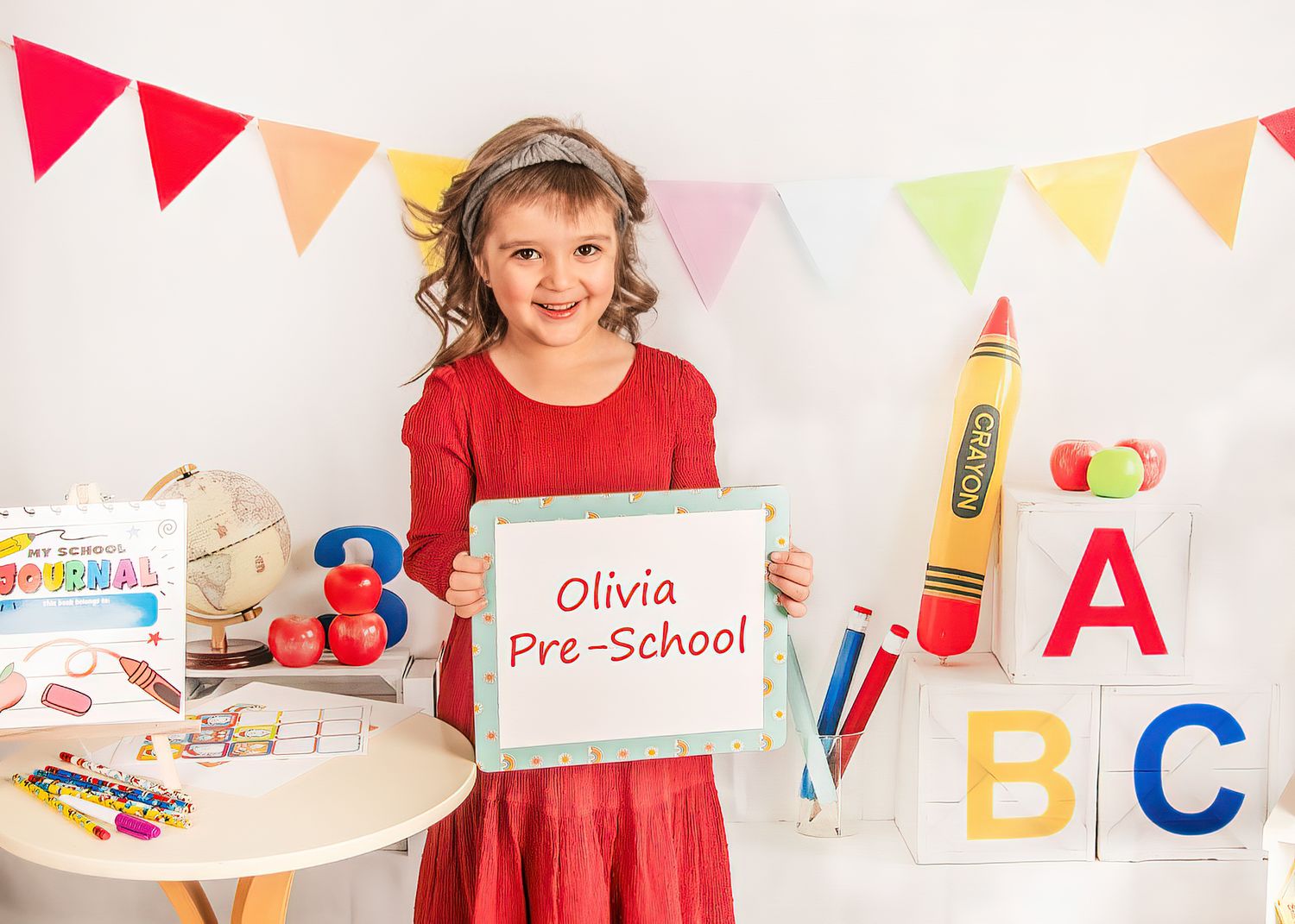 A student in a red dress shows a back-to-school sign at a preschool themed photo session with colorful educational props.