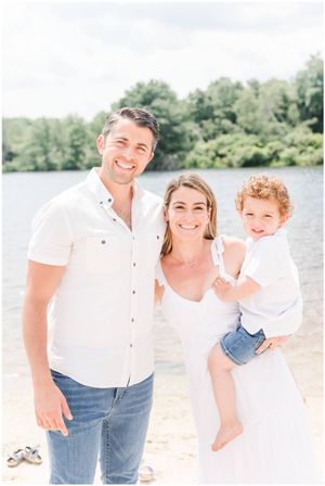 A cheerful group in white attire poses together on a sunny beach with a lake in the background.