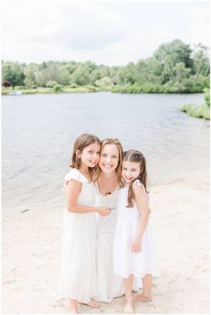 Three figures in white dresses share a joyful moment together on a sandy beach next to a serene lake.