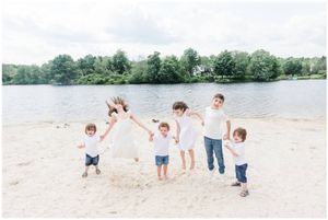 A joyful family wearing white outfits jumps together on a beach by a lake.