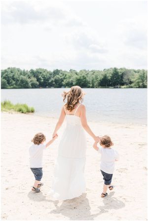 A person in a white dress holds hands with two small children while walking on a sandy beach.