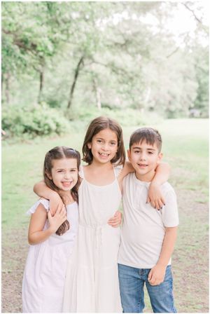 Three children in white and casual outfits pose together outdoors with natural greenery in the background.