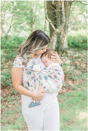 A person wearing a floral print top carries a baby in a patterned wrap carrier outdoors.