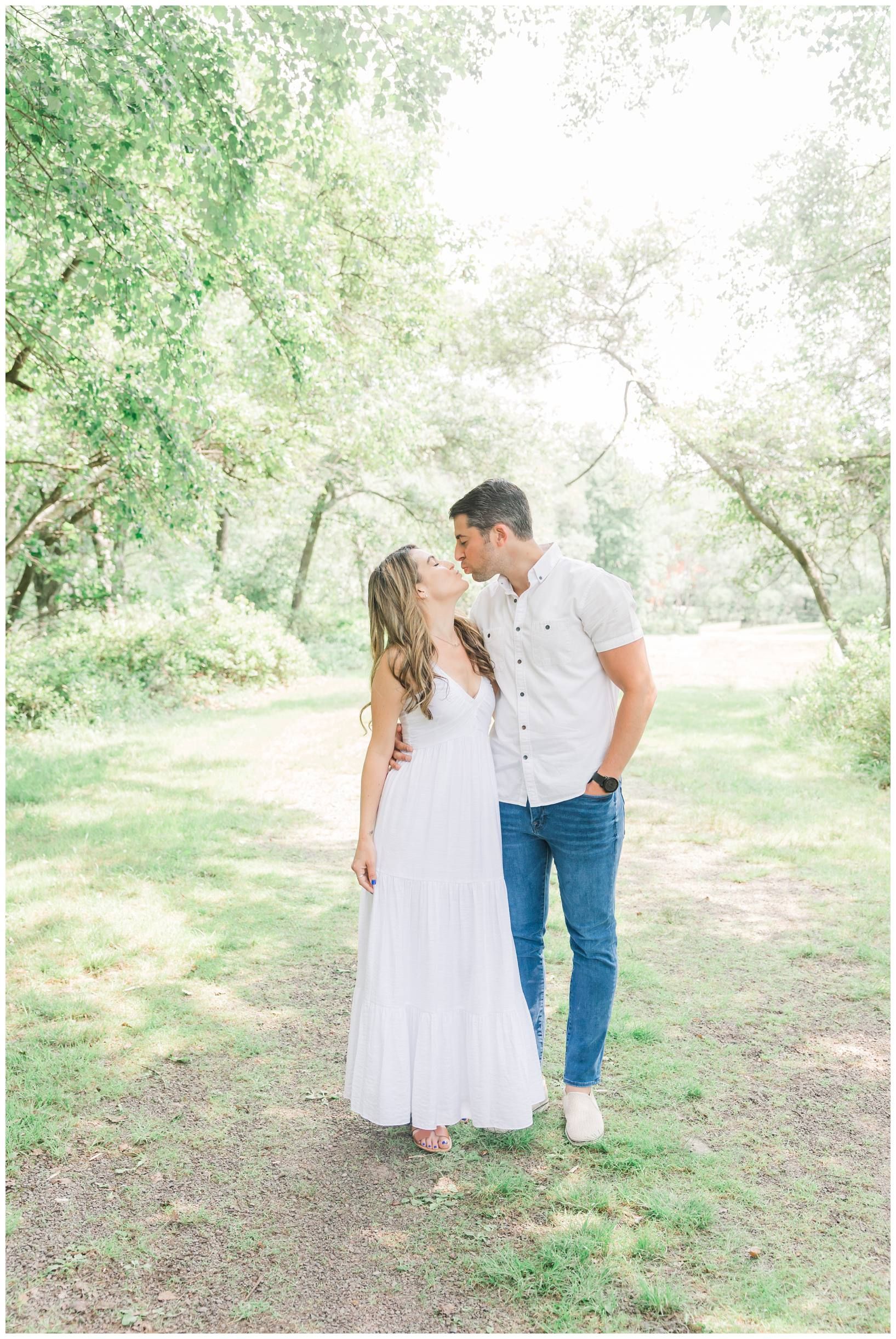 A romantic couple in white and denim outfits stands together in a dreamy outdoor forest setting.