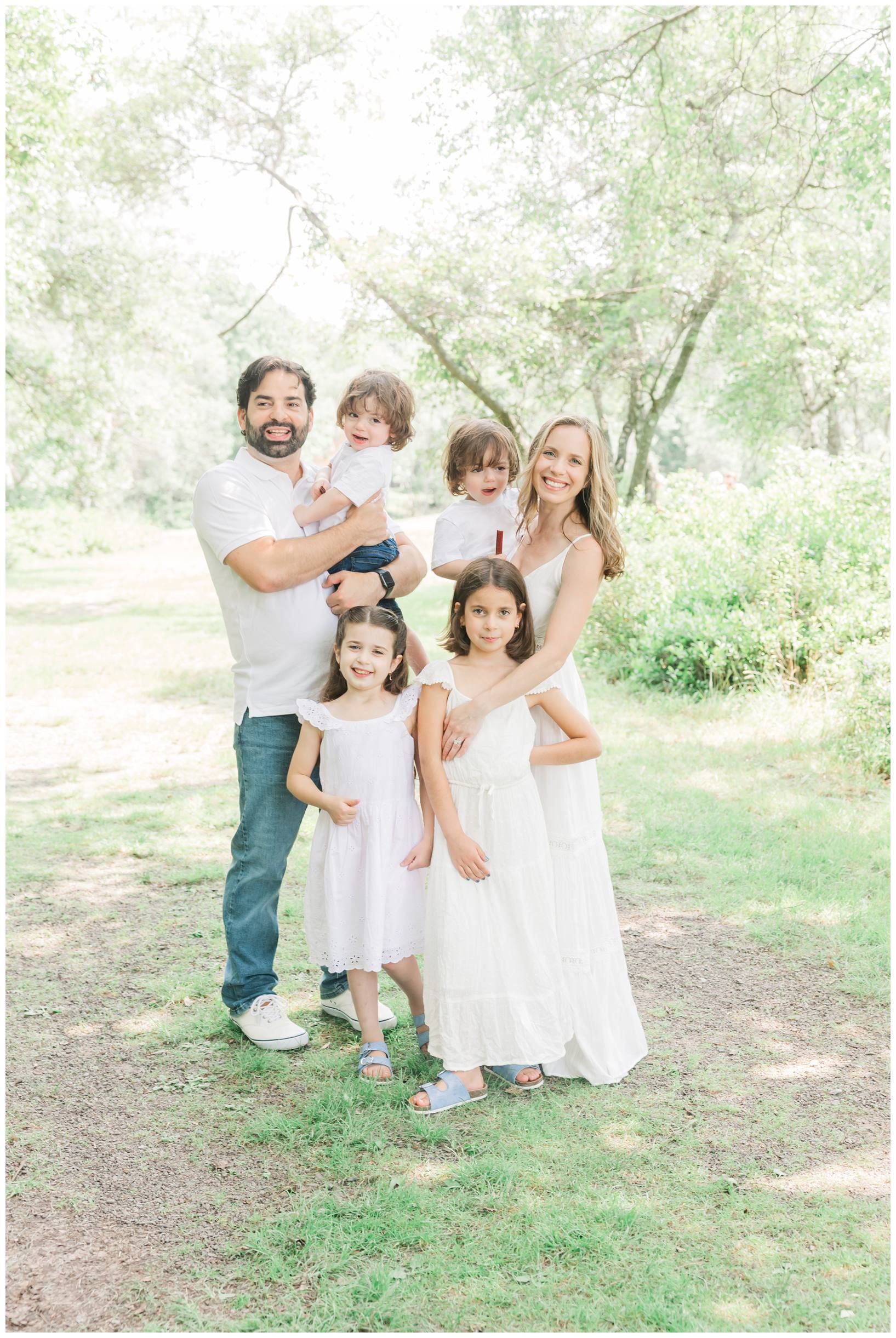 A family of five wearing coordinating white outfits poses together in a sunlit outdoor setting.