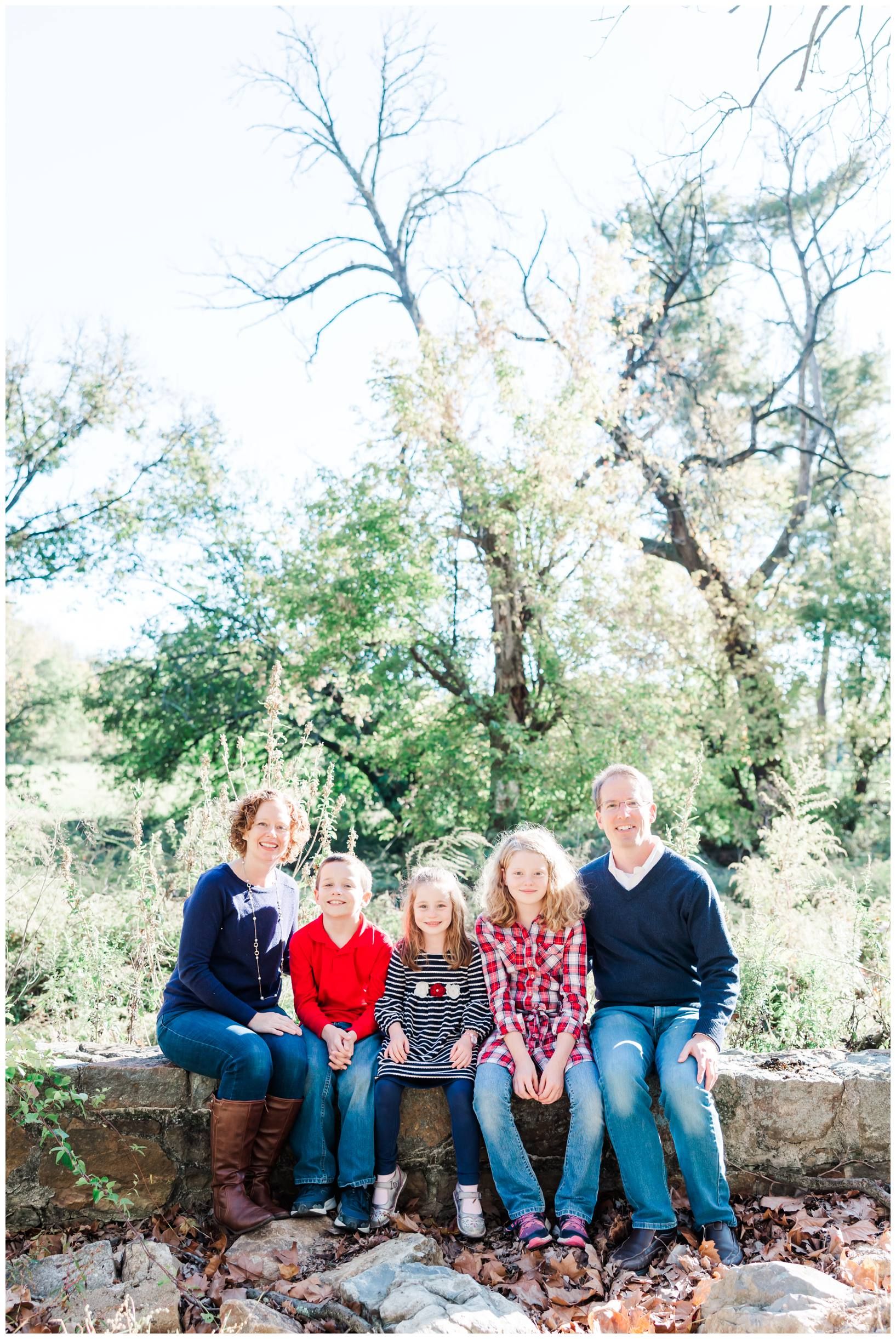 Family of five sits together on rustic stone wall with autumn trees in background.