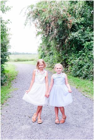 Two children in pretty dresses hold hands while standing on gravel path beside flowering bushes.
