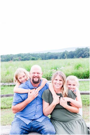 Family of four sits together smiling on wooden fence in green summer field.