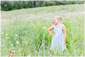 A person in a light blue dress stands among tall grasses and wildflowers in a sunny field.