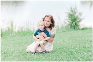 Parent and child share tender moment while sitting on grass near lake.