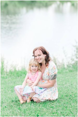 Mother and child sit together on grassy hill by water in summer.