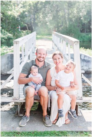 Family of four sits together on white wooden dock over water on sunny day.