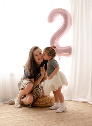 Mother and child share tender moment with pink number 2 balloon in bright room.