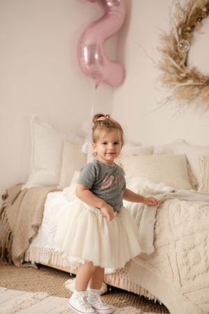 Child in white tulle dress sits on couch with pink balloon decoration.