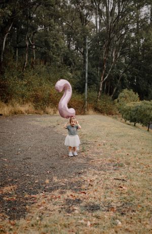Child in white dress plays with pink balloon outdoors in autumn setting.
