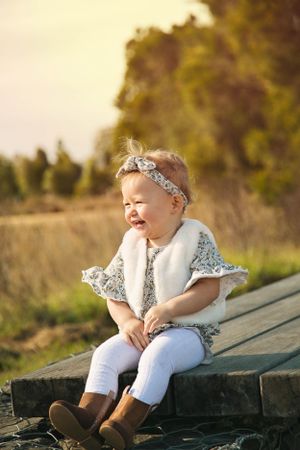 A joyful toddler wearing a floral dress and white vest sits on wooden steps outdoors in warm sunlight.