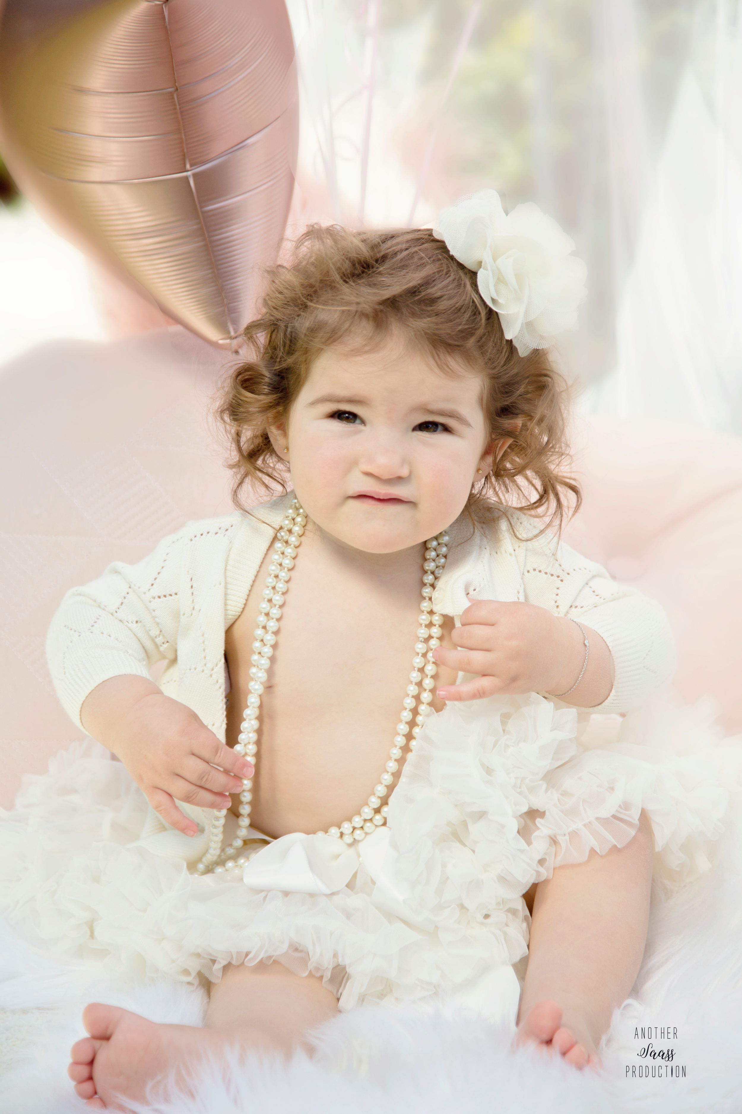 Baby in white lace dress and pearl necklace posing against light background.