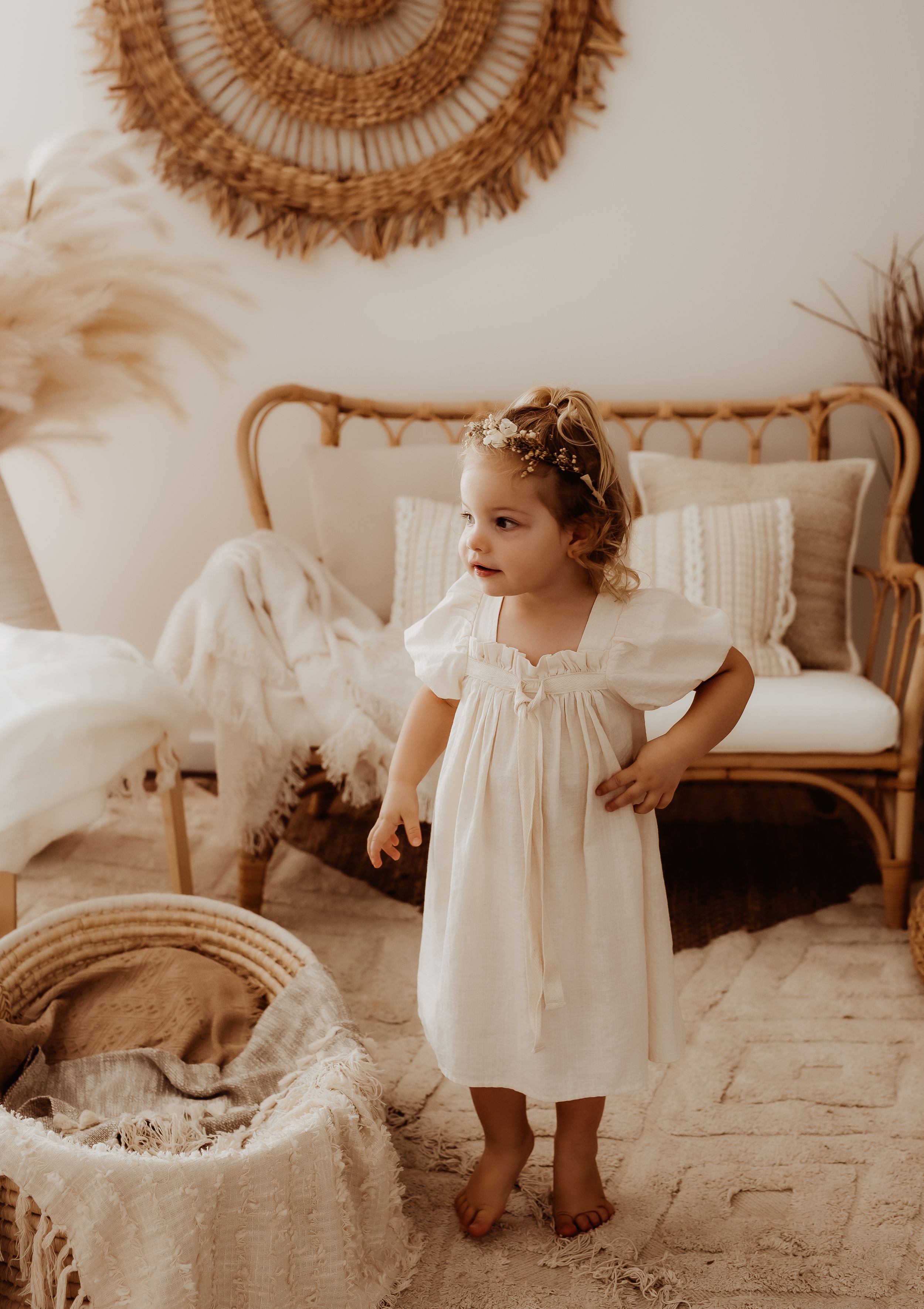 White-dressed child standing near rattan decor in warm, minimalist setting.