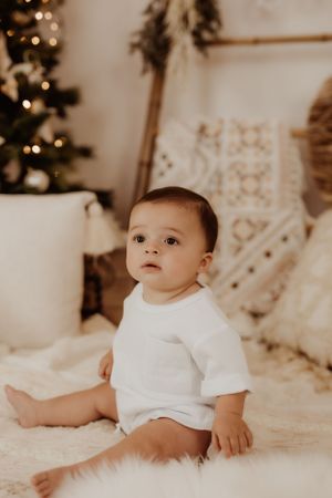 Baby in white onesie sitting on textured bedding with holiday lights in background.