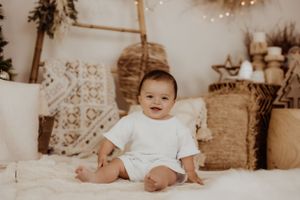 Baby milestone photo with white outfit against neutral holiday backdrop.