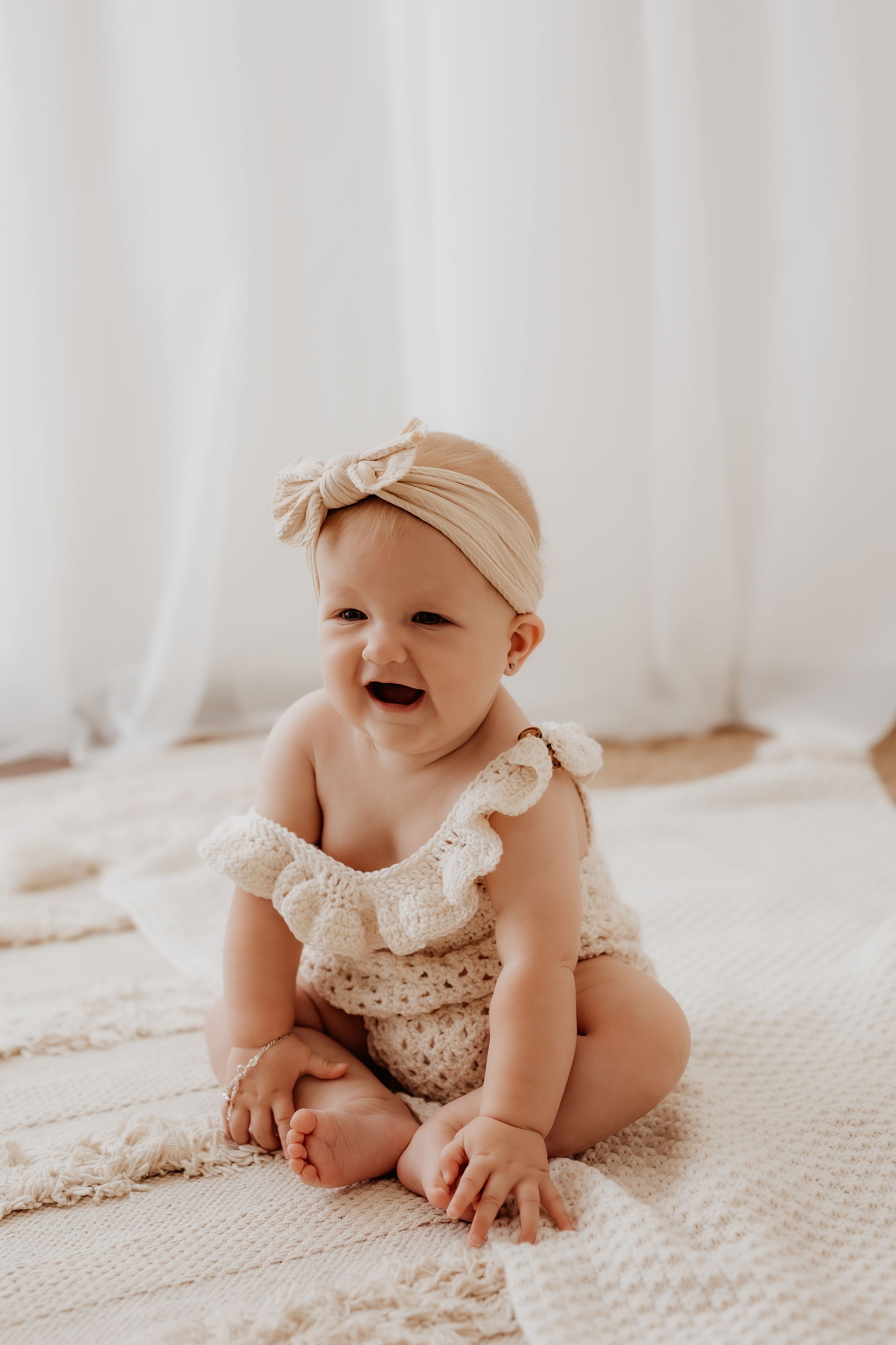 Baby in white lace romper smiles and plays on white bedding during indoor photo session.