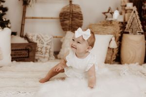 Baby in white dress and bow crawling on textured blanket with rustic decor.
