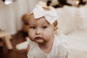 Baby wearing white bow and lace dress looking at camera.
