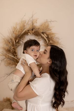 Tender moment between mother and baby against dried floral wreath backdrop.