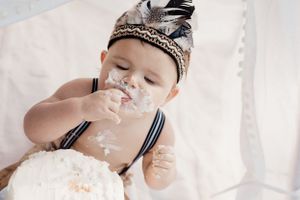Small child wearing a feathered headband messily eating white frosted cake.