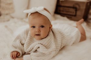 Baby in knit sweater and bow headband lying on white blanket.