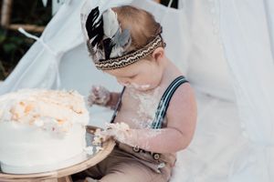 Baby in suspenders and feather crown getting messy with birthday cake.