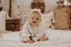 Smiling baby in cream sweater and bow headband sitting on white bedding.