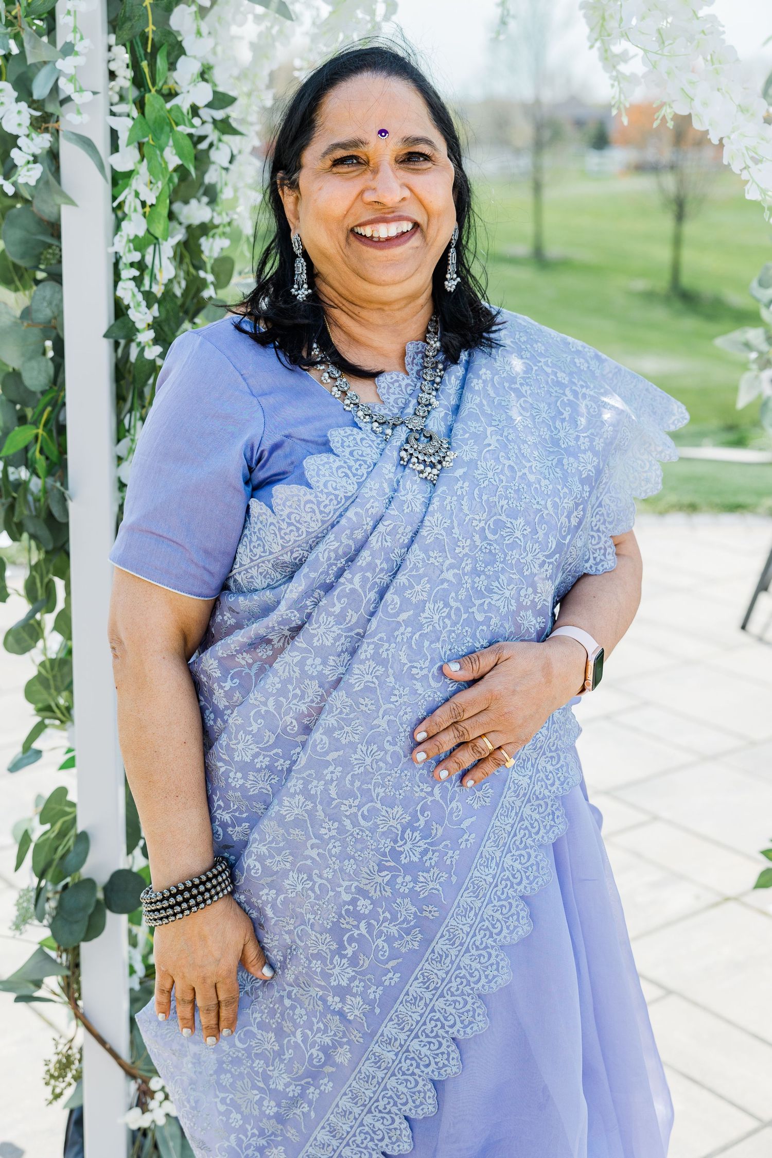 A woman wearing a traditional light blue sari stands smiling under a floral garden arch.