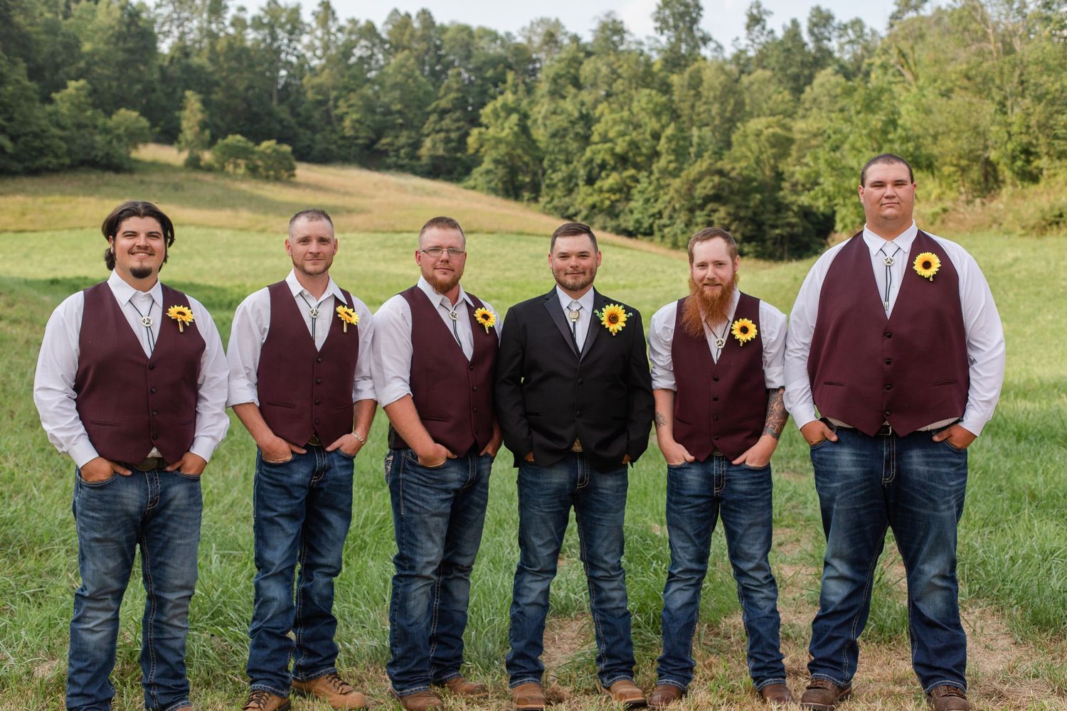 Wedding party members lined up in burgundy vests and jeans on a grassy hillside.