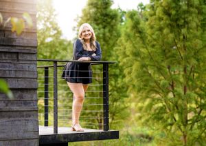 Person in black dress standing on wooden deck railing with green trees in background.